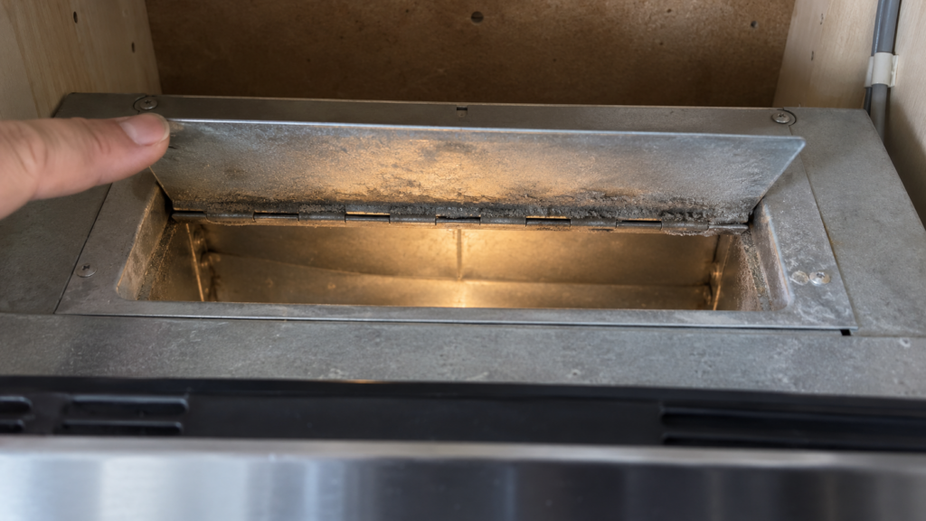 A fingertip holds the metal exhaust damper flap of an over-the-range microwave half open, with grease dust on its hinge.
