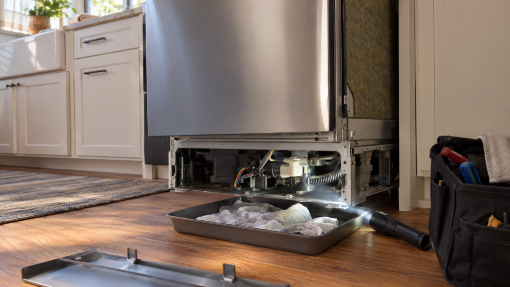 A built-in dishwasher pulled forward out of its cabinet opening with the lower kick plate off, a flashlight beam crossing the exposed lower area and a shallow tray of towels on the kitchen floor, an American home, warm afternoon light.